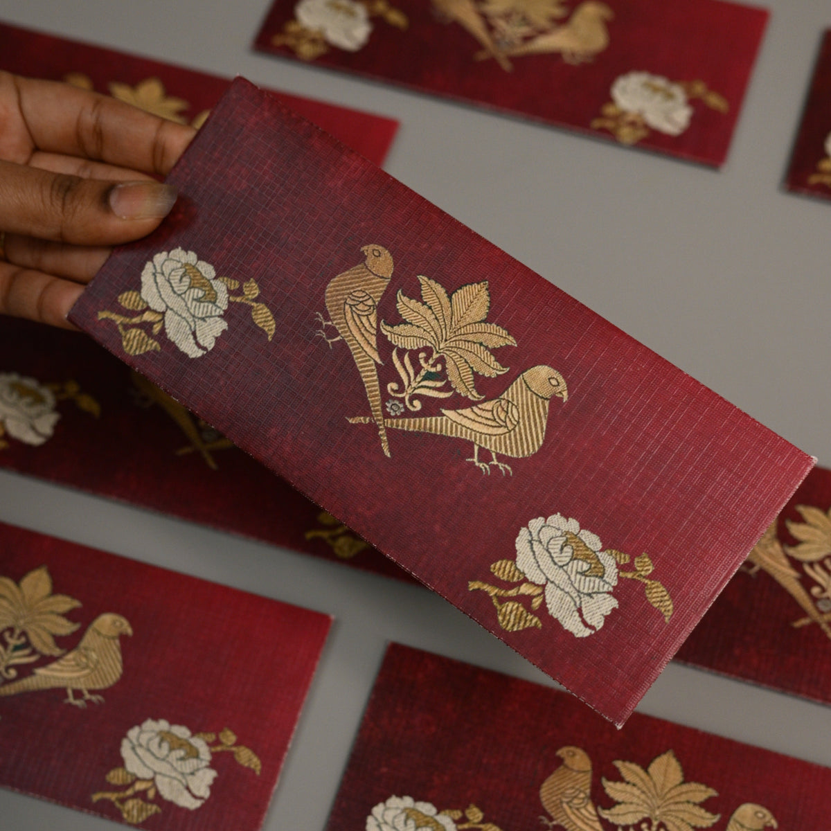 A hand holds a rectangular red fabric card or label featuring a gold bird and floral pattern, with several matching red cards scattered on a gray surface in the background.