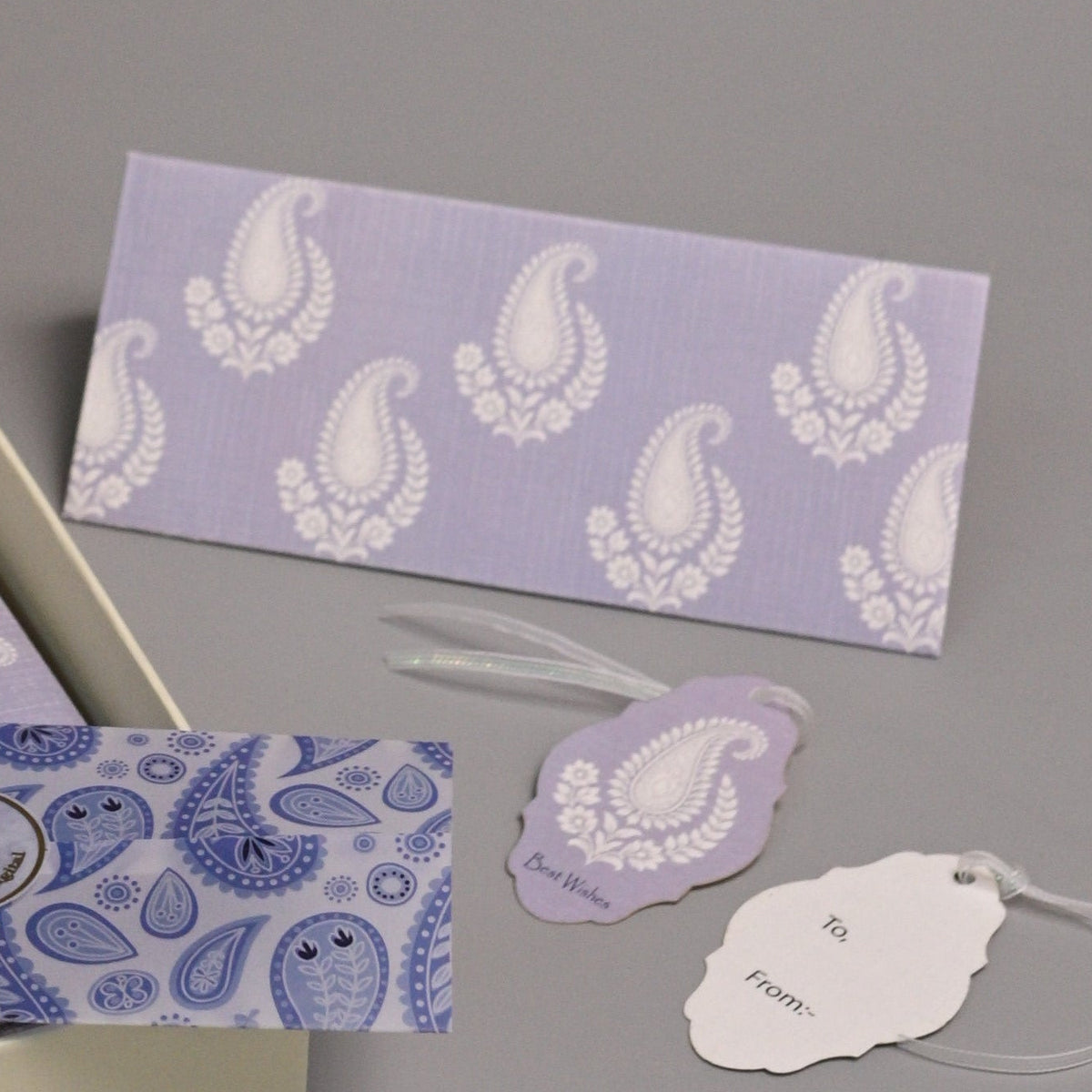 An open decorative gift box with lavender and white patterned items inside, displayed on a gray studio background with matching tags and small cards arranged around it.