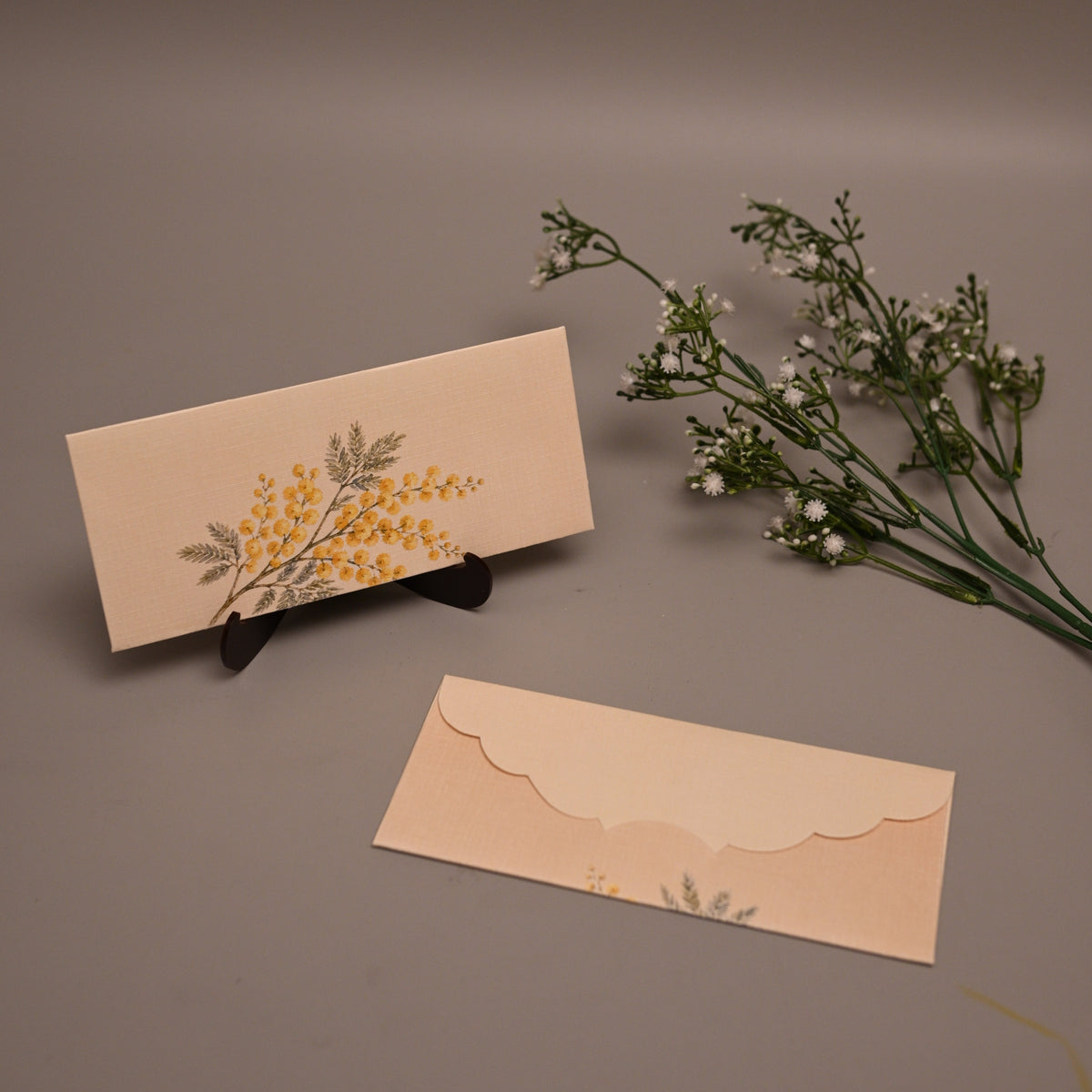 A beige stationery set featuring a rectangular card with a yellow and green floral illustration on a small black stand, and a matching scalloped envelope below, styled with dried baby's breath flowers on a neutral gray background.