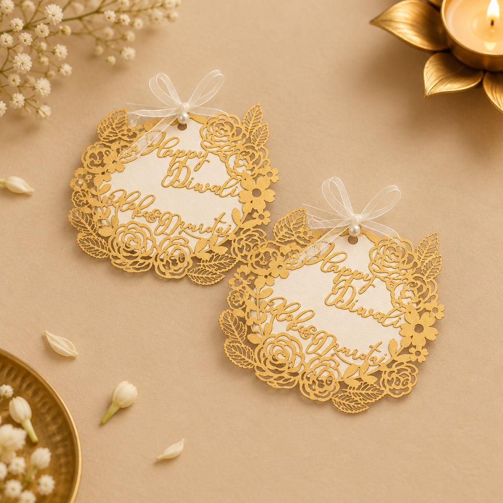 Exquisite laser-cut 'Happy Diwali' gift tags with gold floral and leaf cutouts on a white background, attached with a satin ribbon and a pearl bead.