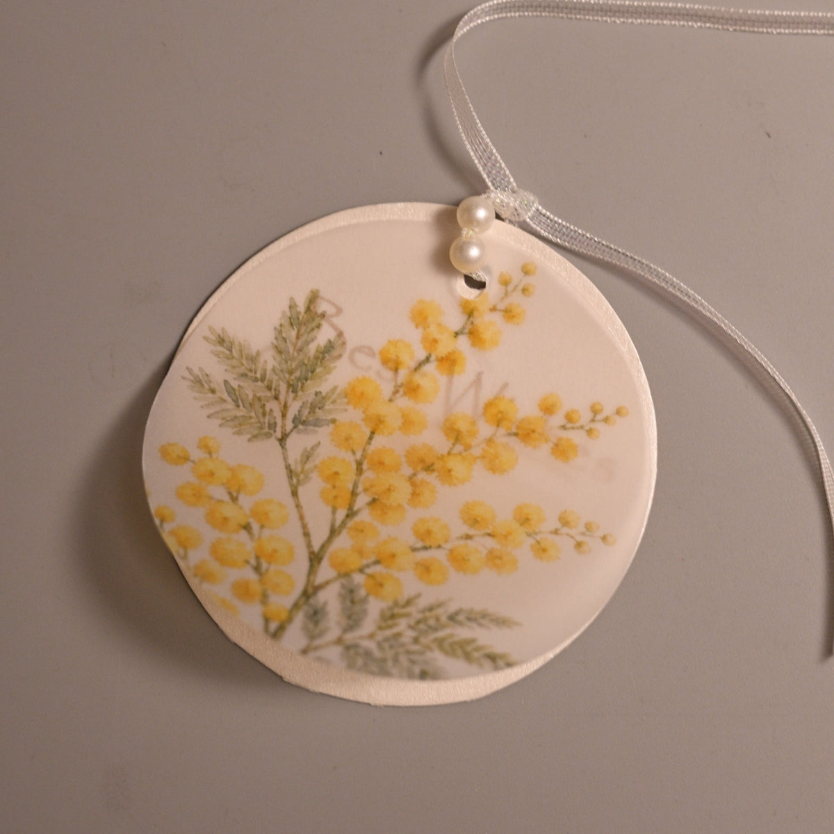 A round butter paper tag with a hand-painted yellow mimosa design, attached to a white cord with a small pearl bead.
