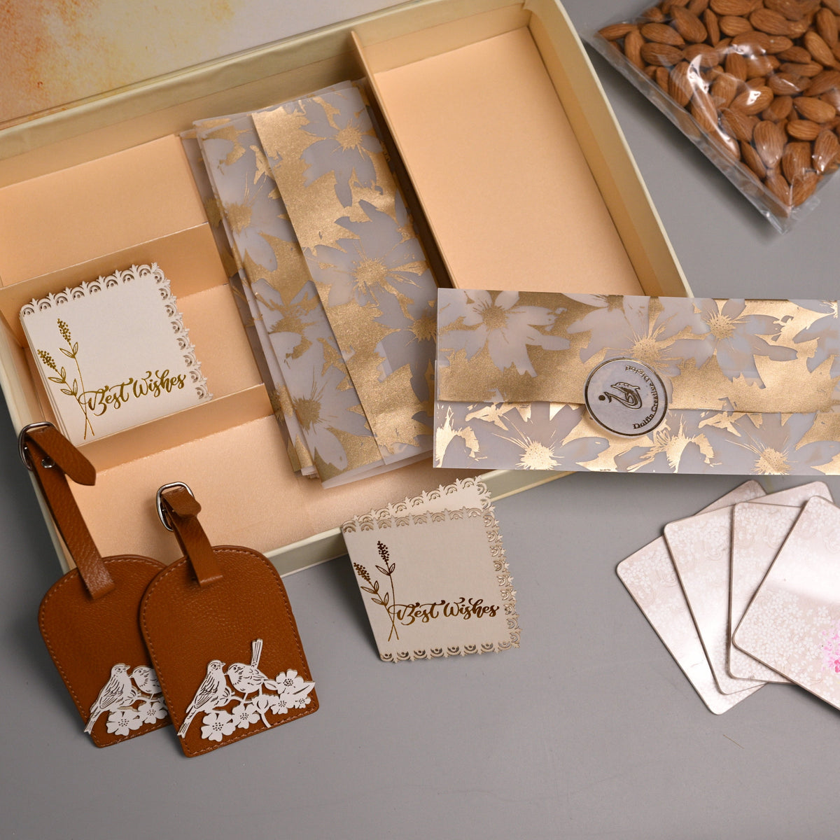 A beige gift box arranged with coordinated stationery patterned envelopes, small cards, a pair of brown leather luggage tags, and a bag of almonds. The set has warm beige, gold, and brown tones