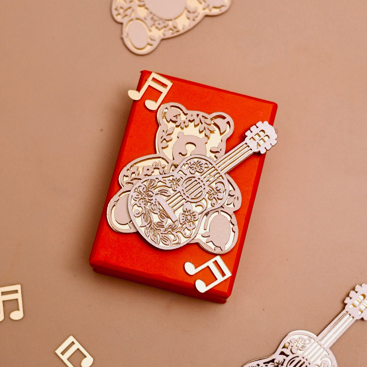 A red rectangular gift box wrapped with a golden acrylic bear with guitar ornament with decorative musical note cutouts.