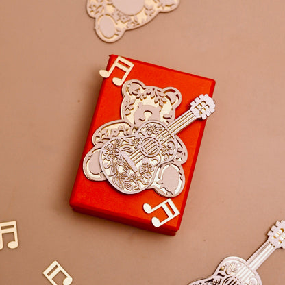 A red rectangular gift box wrapped with a golden acrylic bear with guitar ornament with decorative musical note cutouts.
