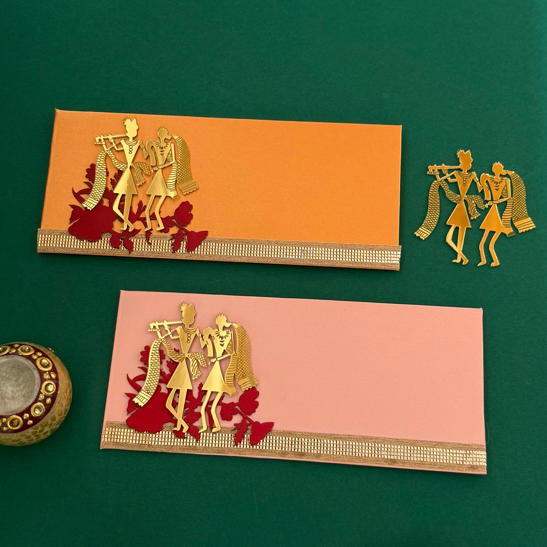 A set of two acrylic envelopes with gold-colored Radha Krishna figures on them, one orange and one pink background, placed on a green surface with a decorative item to the side.
