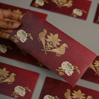 A hand holds a rectangular red fabric card or label featuring a gold bird and floral pattern, with several matching red cards scattered on a gray surface in the background.