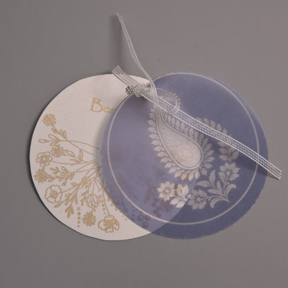 Two round decorative tags or labels with floral and paisley designs, one in white with gold botanical print and the other in translucent lavender with white paisley, tied together with a sheer white ribbon on a neutral gray background.