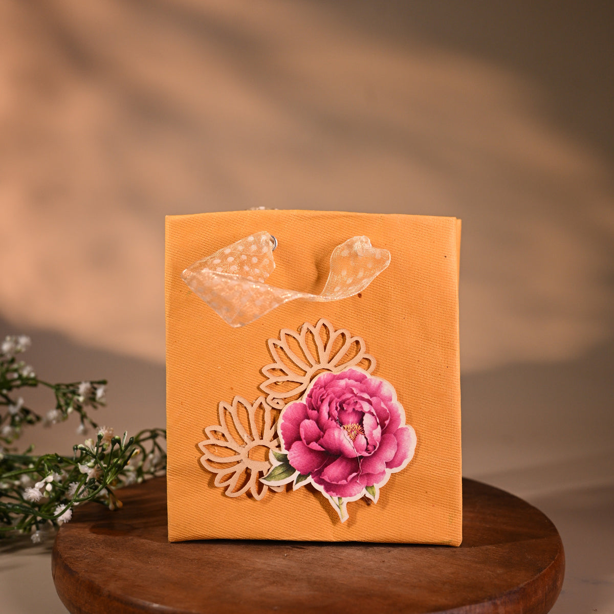 This is a pretty gift bag made from strong paper. It has a beautiful golden floral design with pink flower sticker on the front. The bag has gold ribbon handles, making it easy to carry and giving it a stylish touch.