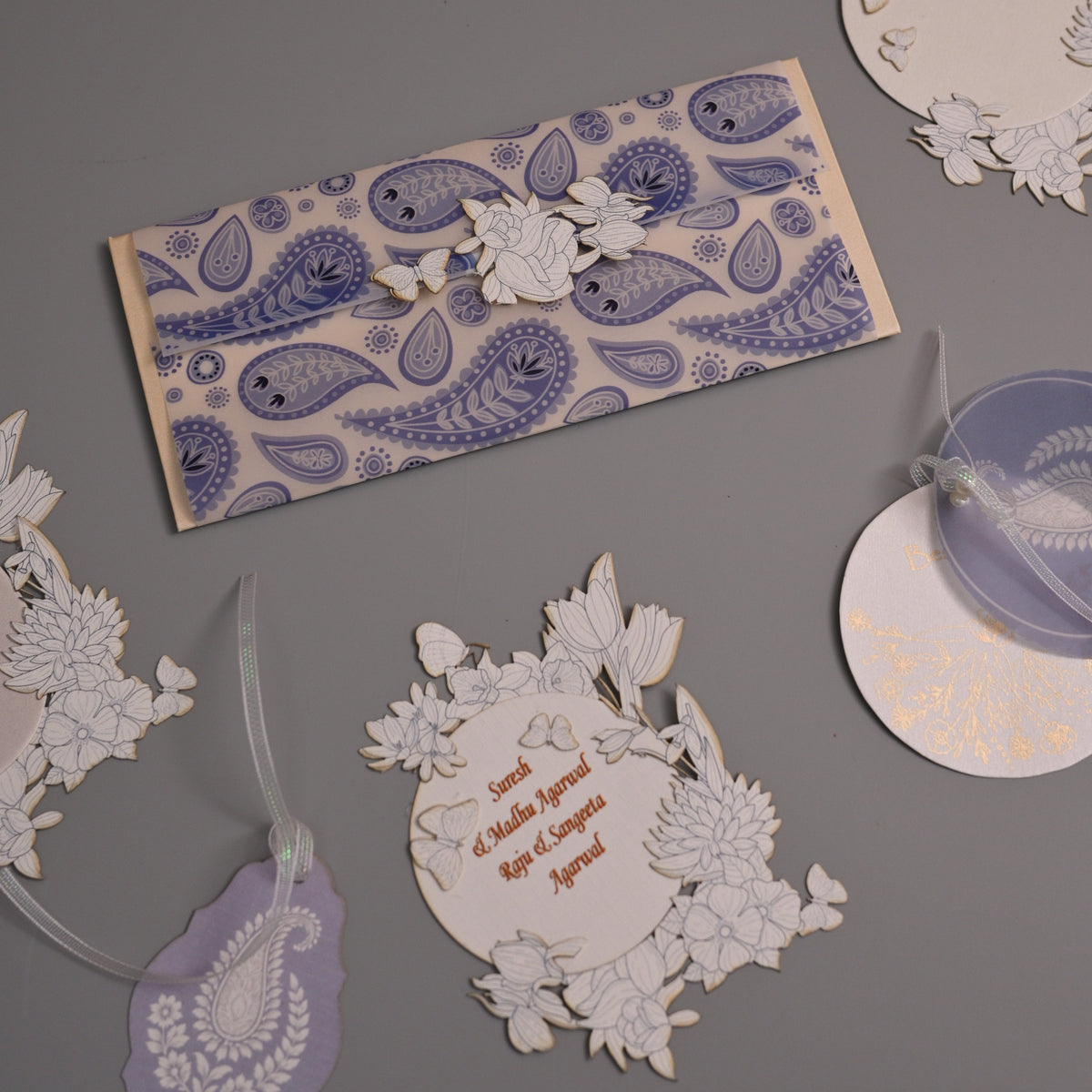 A styled flat-lay of intricate white paper cutouts and tags with blue and purple paisley patterns, arranged on a solid gray background.