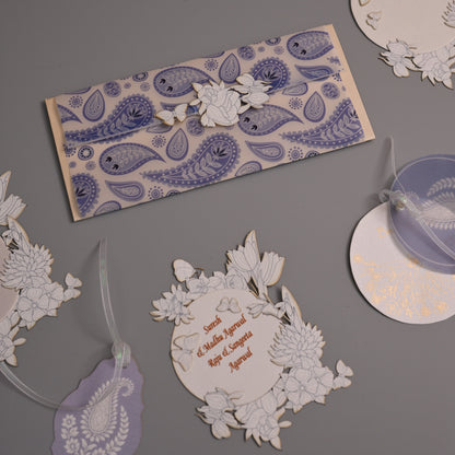 A styled flat-lay of intricate white paper cutouts and tags with blue and purple paisley patterns, arranged on a solid gray background.