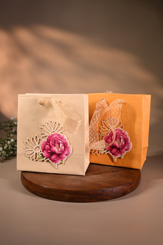 This is a pretty gift bag made from strong paper. It has a beautiful golden floral design with pink flower sticker on the front. The bag has gold ribbon handles, making it easy to carry and giving it a stylish touch.