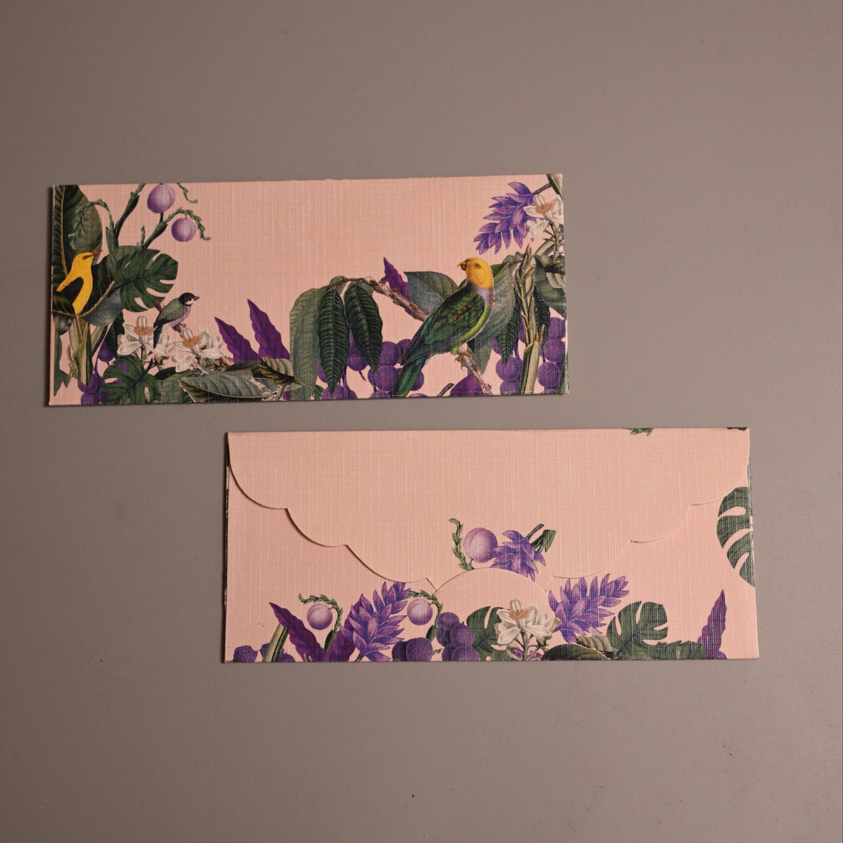 Two rectangular envelopes with a pink background and a tropical bird and floral print, displayed on a plain gray surface.