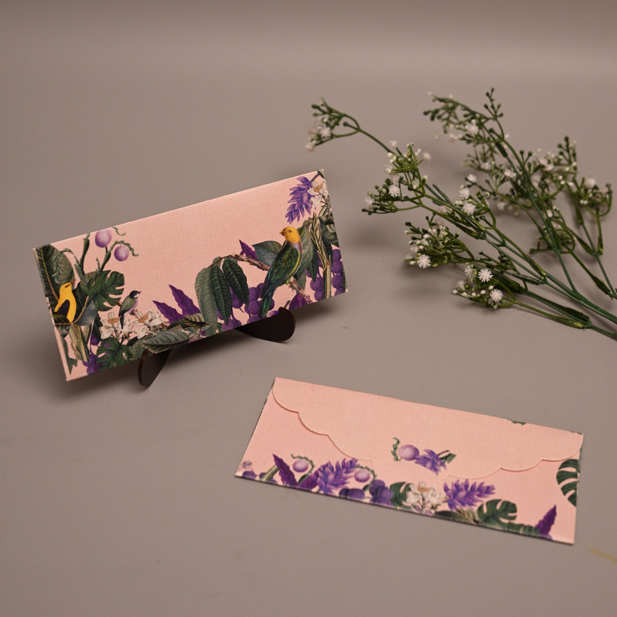 A pink floral clutch purse and matching flat card or envelope set with purple flowers, green leaves, and butterfly motifs, styled on a neutral gray background with small white flowers.