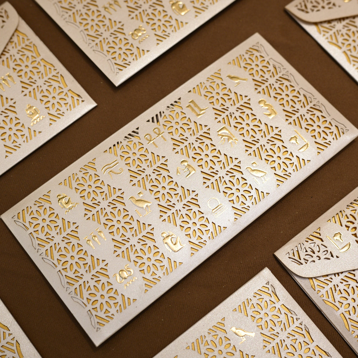 White paper invitation cards and matching envelopes with intricate laser-cut geometric patterns and gold foil lettering, arranged diagonally on a dark brown background.