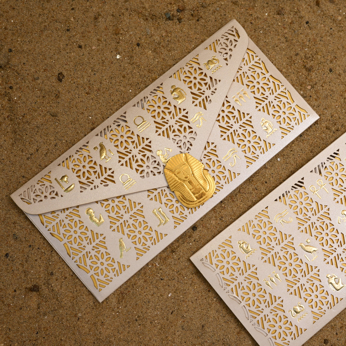 Two white paper envelopes with intricate gold laser-cut patterns and small gold foil details, one closed with a gold clasp and one open, displayed on a sandy brown background.