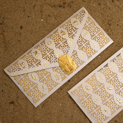 Two white paper envelopes with intricate gold laser-cut patterns and small gold foil details, one closed with a gold clasp and one open, displayed on a sandy brown background.