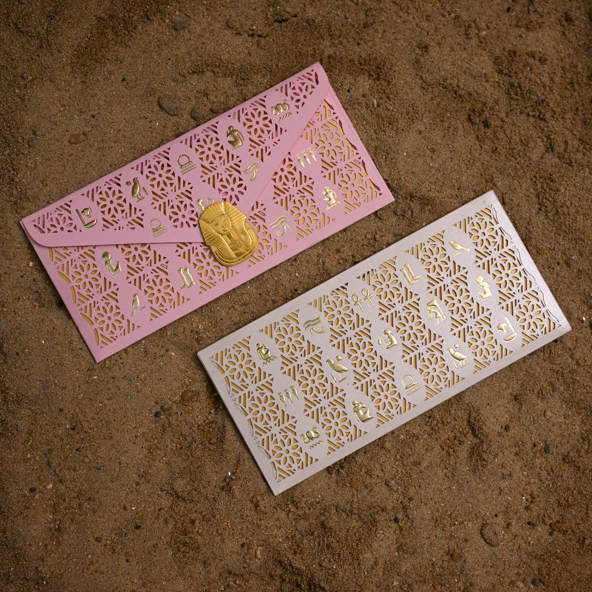 Two laser-cut envelope-style cards or envelopes with intricate gold patterns, one pink and one white, placed on a brown textured surface.