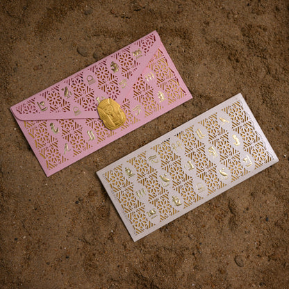 Two laser-cut envelope-style cards or envelopes with intricate gold patterns, one pink and one white, placed on a brown textured surface.