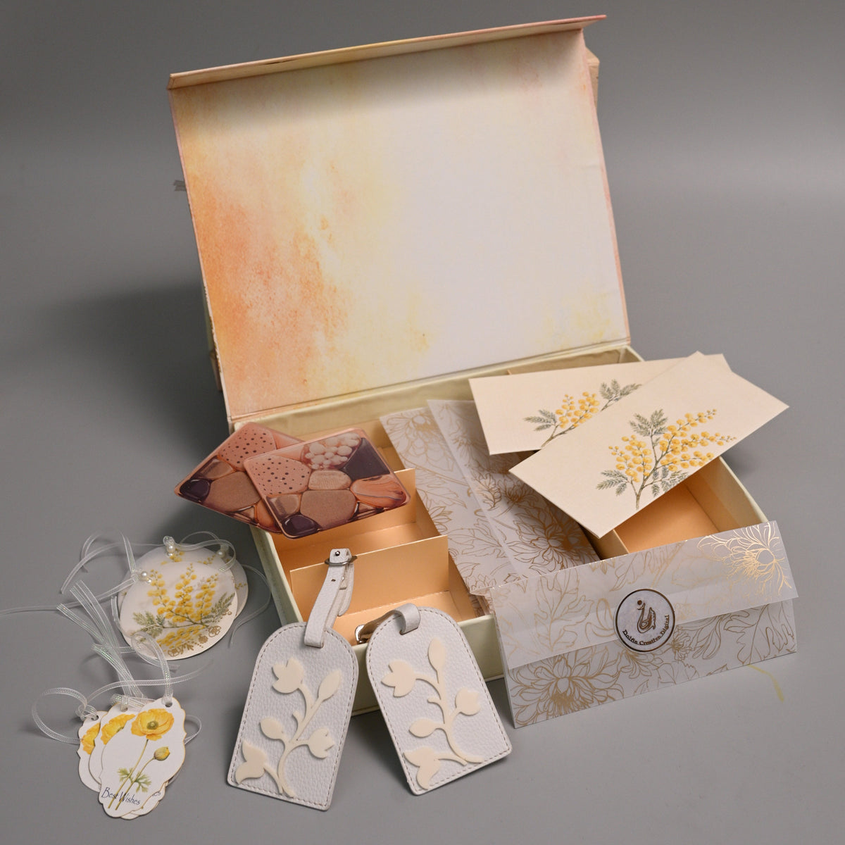 A gift set in an open vintage-style box with a watercolor-style beige and orange interior, containing cream-colored paper tags with yellow floral designs, a small card, and a patterned ribbon, arranged on a gray background.