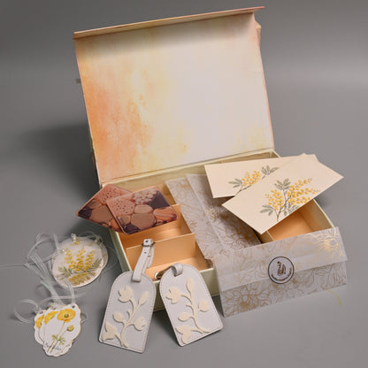 A gift set in an open vintage-style box with a watercolor-style beige and orange interior, containing cream-colored paper tags with yellow floral designs, a small card, and a patterned ribbon, arranged on a gray background.