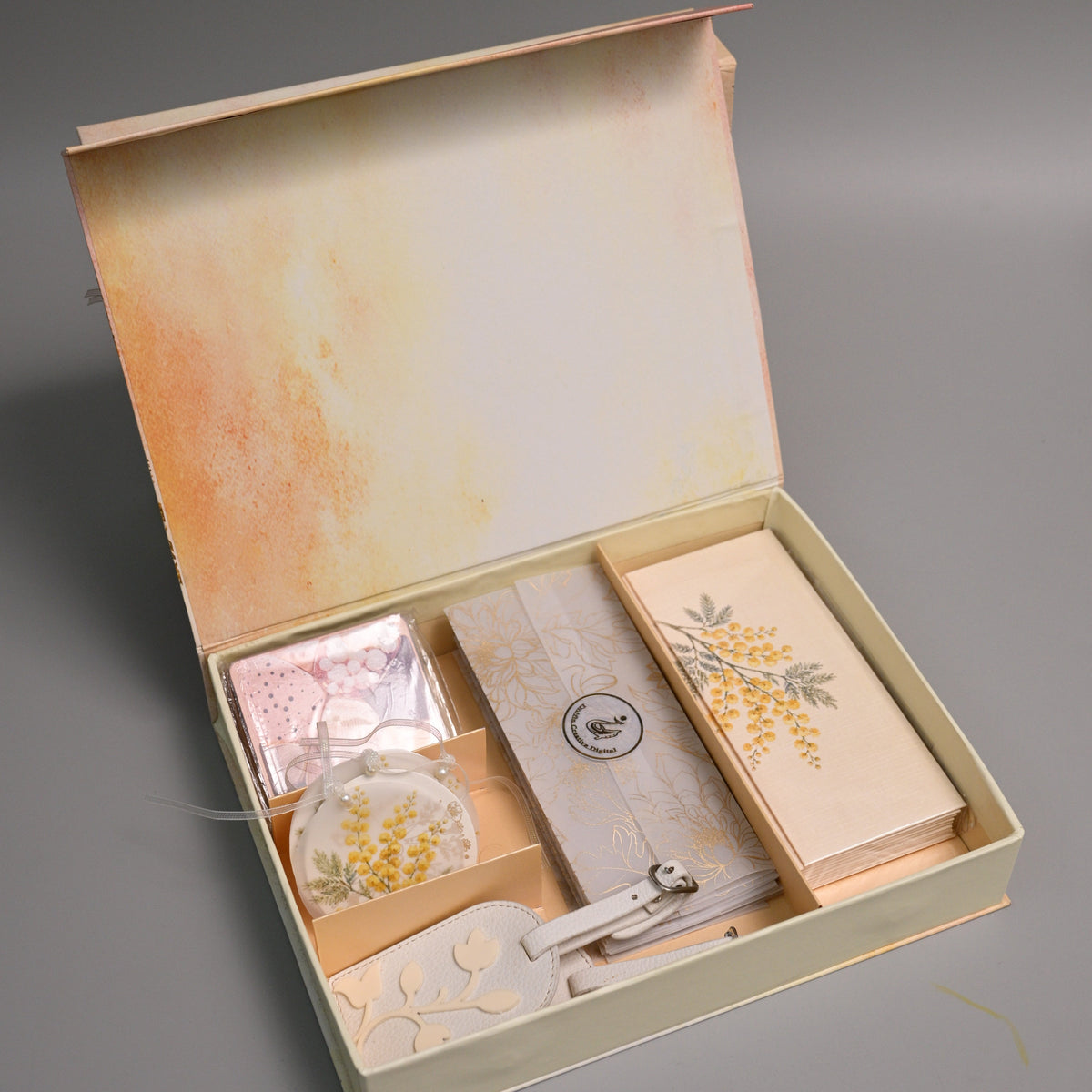 An open cream-colored gift box with a warm orange-brown interior, containing a small pink wrapped item, a rectangular beige card or book with yellow floral artwork, a small round object, and a white decorative piece, all photographed on a neutral gray background.