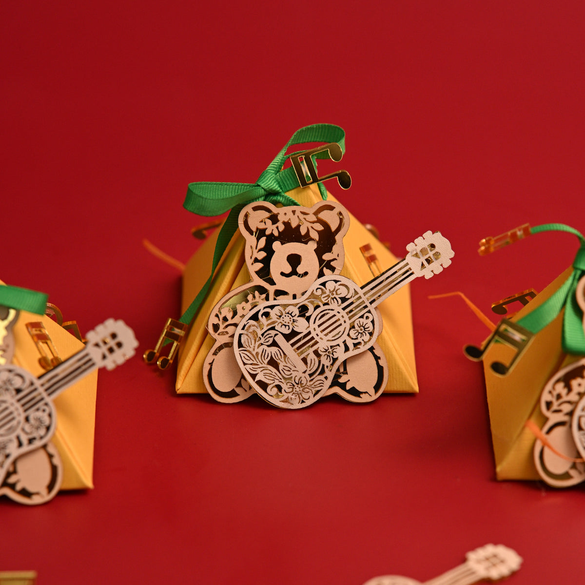 Three small triangular gift boxes in warm yellow are arranged on a solid red background, each decorated with a green ribbon and a laser-cut teddy bear holding an acoustic guitar. A loose laser-cut guitar decoration lies in the foreground.