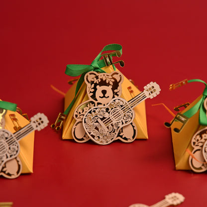 Three small triangular gift boxes in warm yellow are arranged on a solid red background, each decorated with a green ribbon and a laser-cut teddy bear holding an acoustic guitar. A loose laser-cut guitar decoration lies in the foreground.