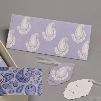 An open decorative gift box with lavender and white patterned items inside, displayed on a gray studio background with matching tags and small cards arranged around it.