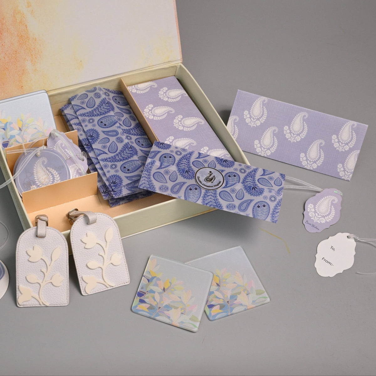 A gift set containing lavender and blue paisley patterned cards, envelopes, and decorative tags, displayed in an open cream-colored box on a gray surface.
