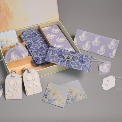 A gift set containing lavender and blue paisley patterned cards, envelopes, and decorative tags, displayed in an open cream-colored box on a gray surface.