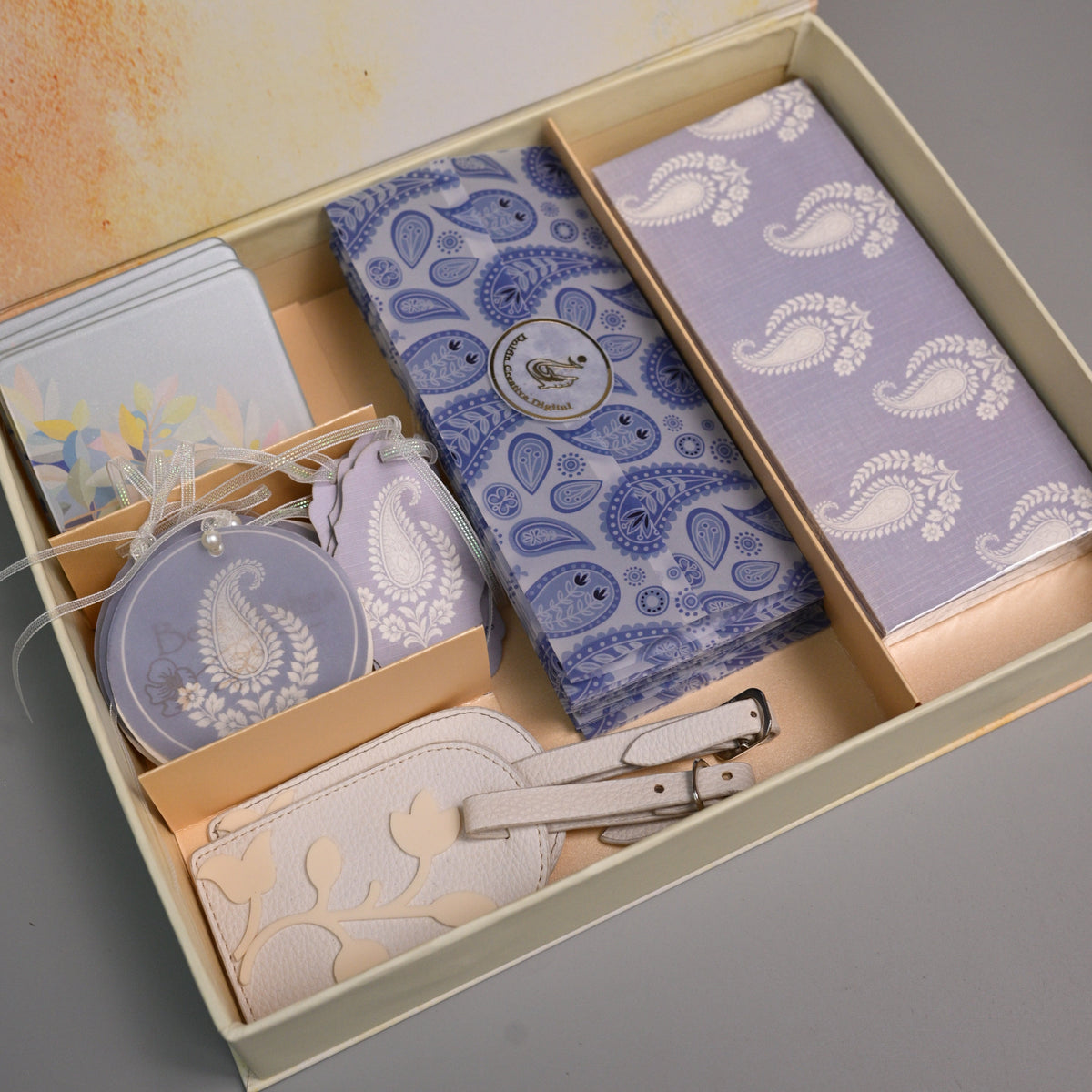 A gift set in a beige box with a watercolor-style printed interior lid, containing a blue paisley-patterned wallet, a lavender paisley-patterned card holder, a matching zippered pouch, a small round tag, and a cream-colored leather strap.