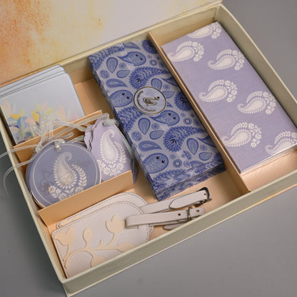 A gift set in a beige box with a watercolor-style printed interior lid, containing a blue paisley-patterned wallet, a lavender paisley-patterned card holder, a matching zippered pouch, a small round tag, and a cream-colored leather strap.