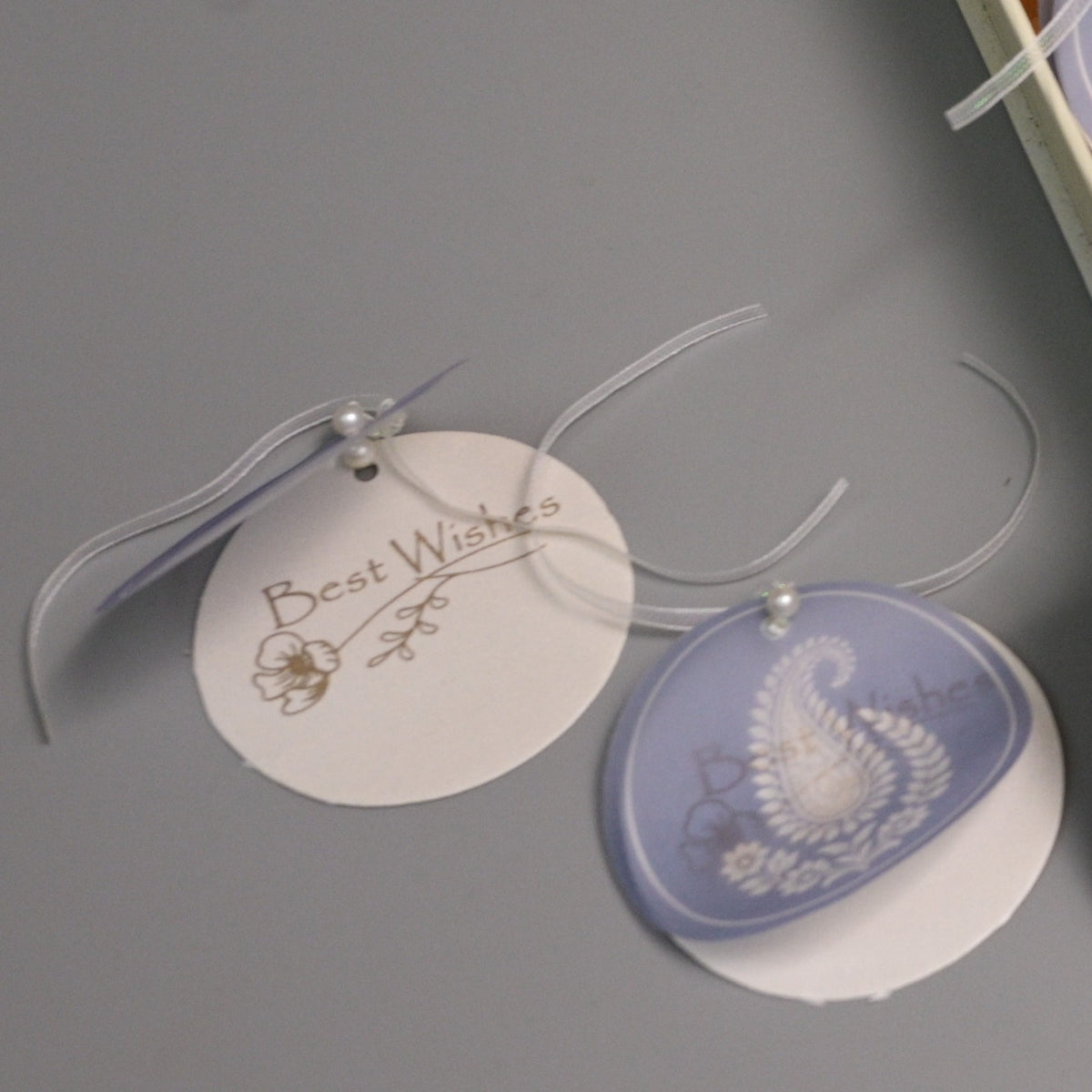 Two round hanging gift tags on clear ribbons, one white with gold script reading 'Best Wishes' and one pale lavender with a white paselay 