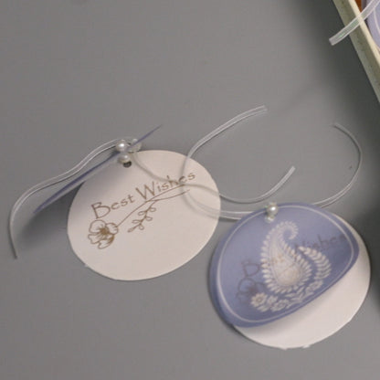 Two round hanging gift tags on clear ribbons, one white with gold script reading 'Best Wishes' and one pale lavender with a white paselay 