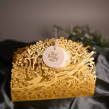 A yellow laser-cut paper envelope or box with intricate floral and branch patterns, featuring a small round white hanging tag