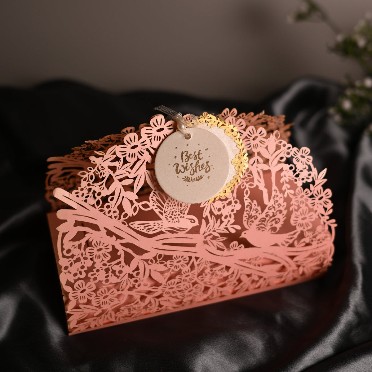A rose gold laser-cut decorative box with intricate floral and bird cutout patterns, featuring a hanging round tag that reads 'Best Wishes' in gold lettering