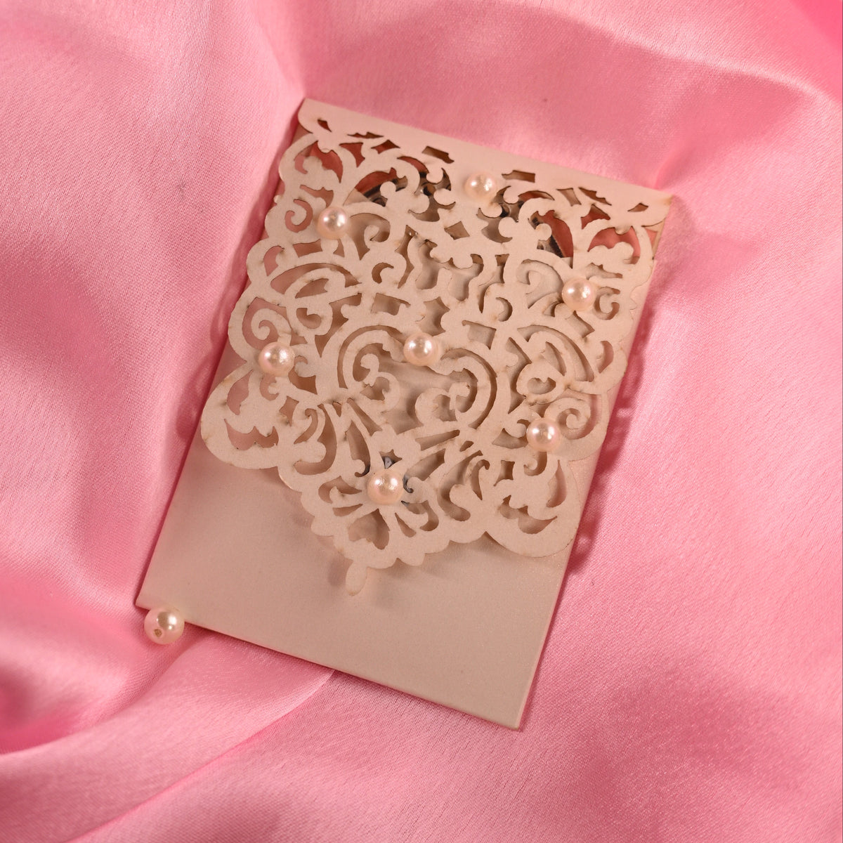 A beige rectangular card or envelope with an ornate laser-cut floral pattern and small pearl embellishments, placed on draped pink satin fabric.