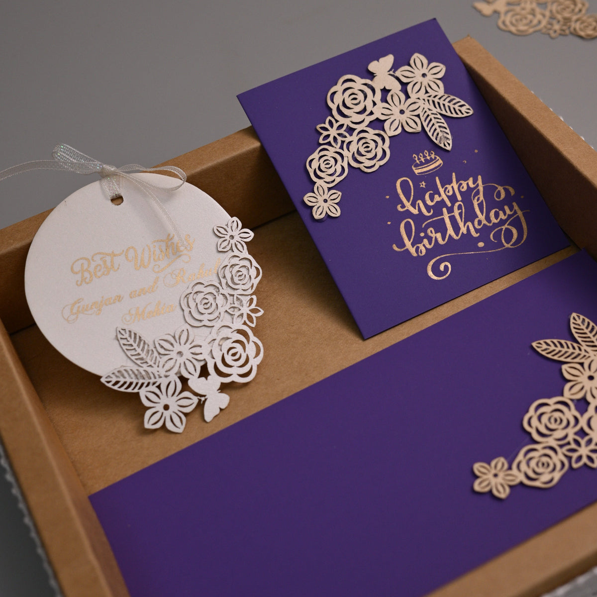 A luxury laser-cut stationery set in a kraft gift box, featuring a purple envelope with gold 'Happy Birthday' script, a matching round white paper tag, and a purple card, all decorated with intricate white floral laser-cut borders