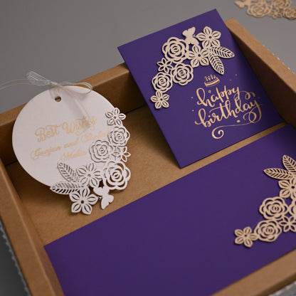 A luxury laser-cut stationery set in a kraft gift box, featuring a purple envelope with gold 'Happy Birthday' script, a matching round white paper tag, and a purple card, all decorated with intricate white floral laser-cut borders