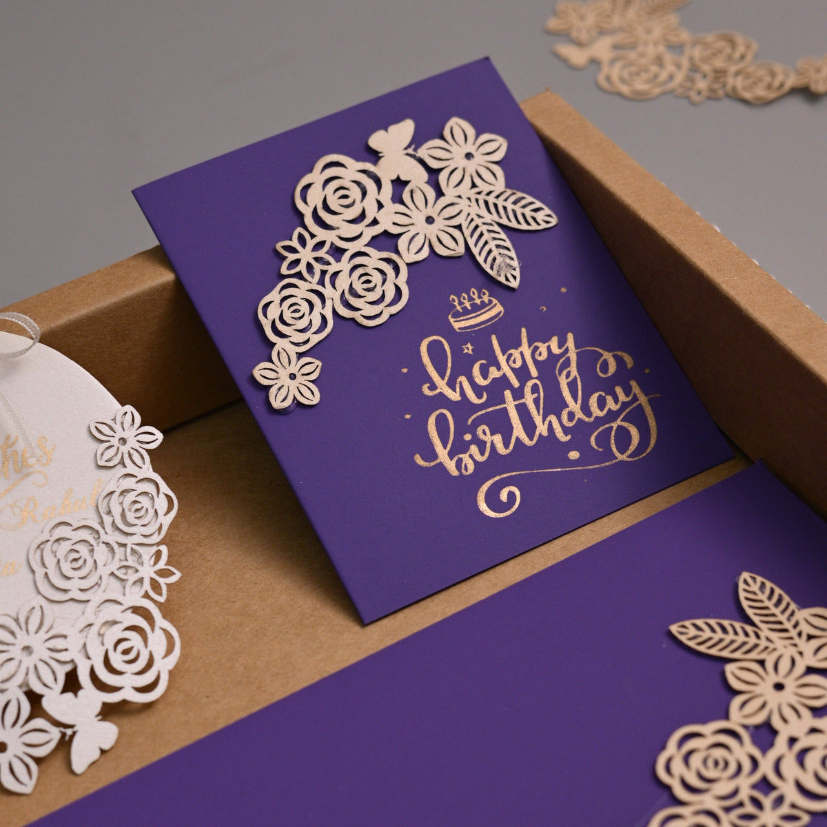 A luxury laser-cut paper stationery set in a kraft cardboard box, featuring a purple card with gold foil script reading 'Happy Birthday', a matching purple envelope, and a white circular gift tag with laser-cut floral details.