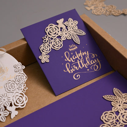 A luxury laser-cut paper stationery set in a kraft cardboard box, featuring a purple card with gold foil script reading 'Happy Birthday', a matching purple envelope, and a white circular gift tag with laser-cut floral details.
