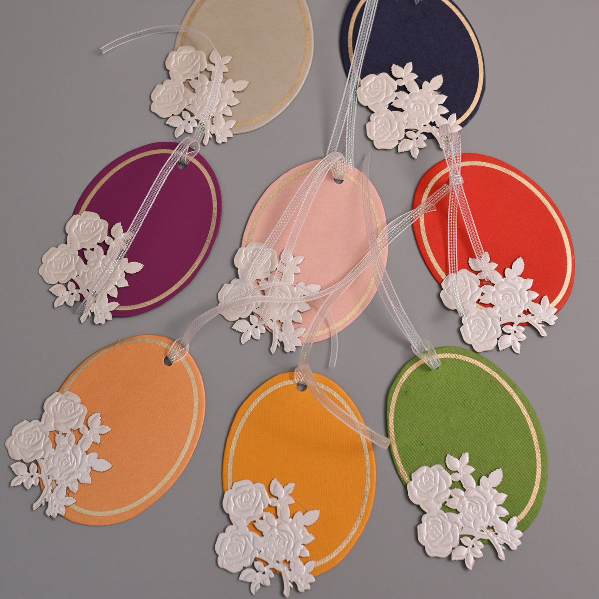 A set of colorful oval paper tags with white floral cutout embellishments, hanging from white ribbons.