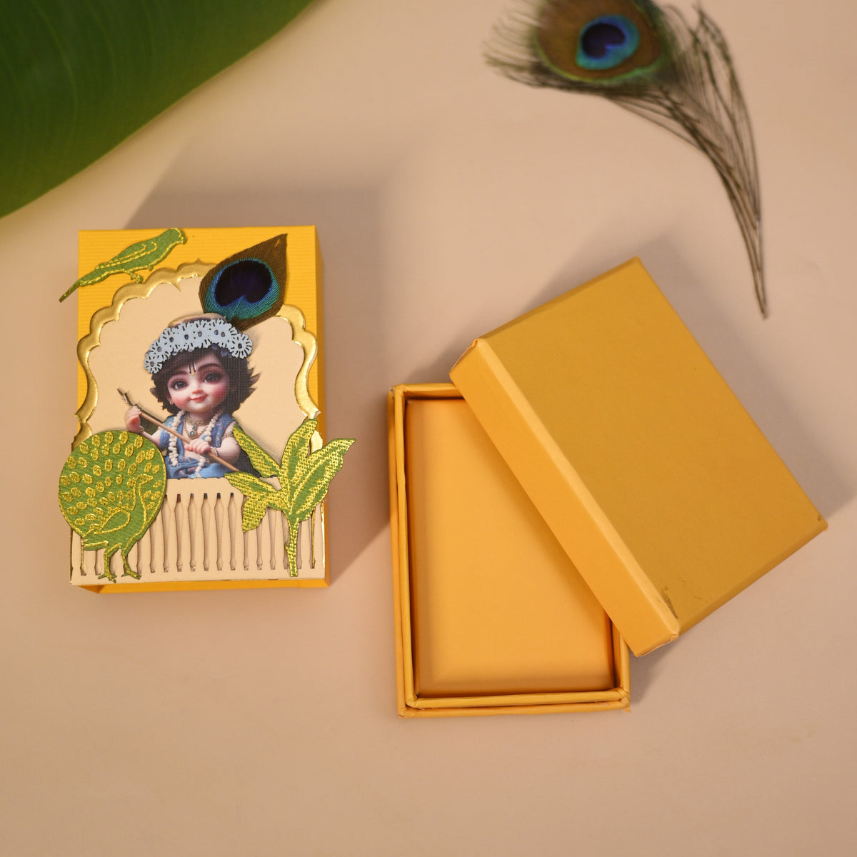 A mustard yellow decorative coin box with a foil embossed Krishana and peacock feather motif, shown next to an open matching empty box