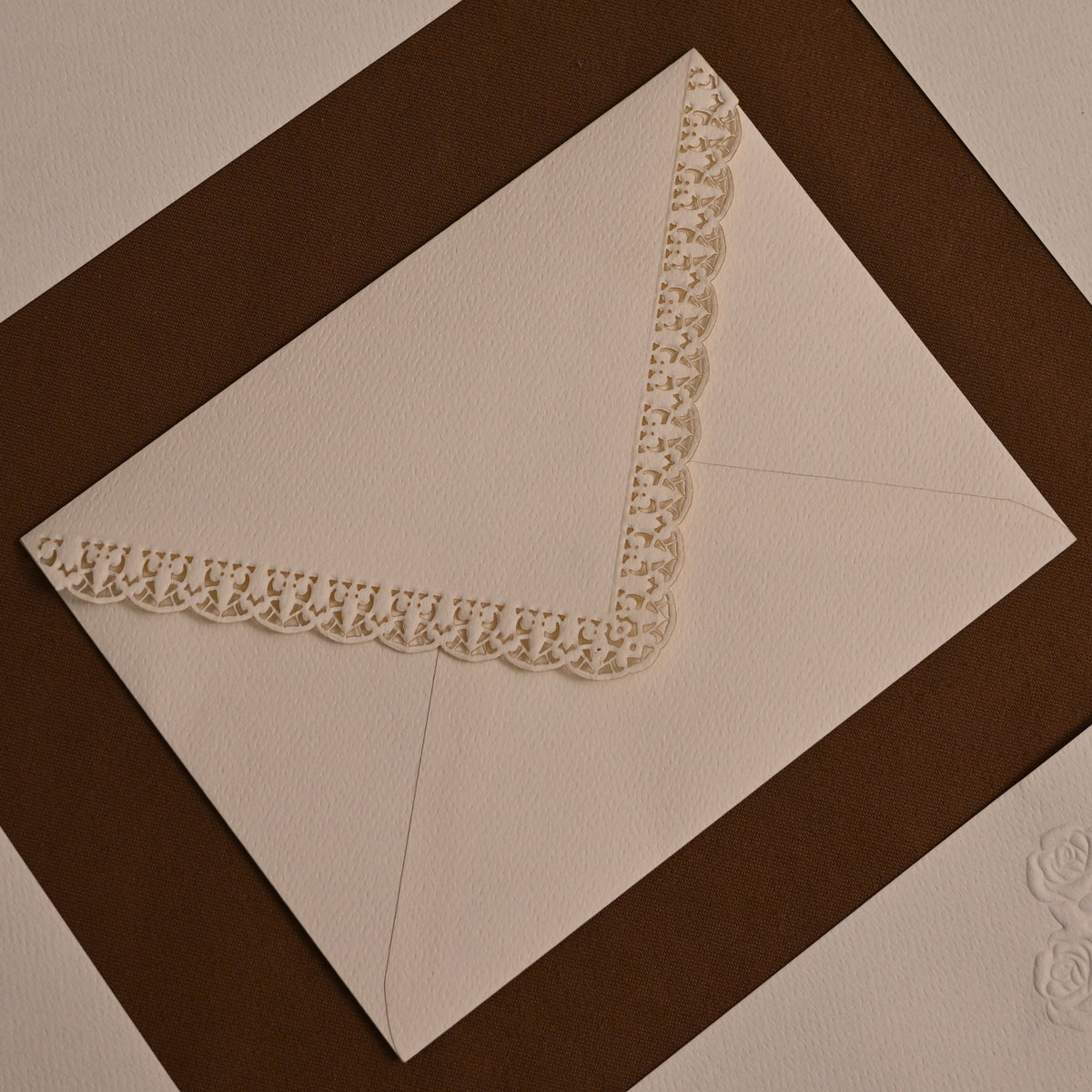A close-up of cream-colored paper envelopes arranged in a geometric pattern with dark brown borders. One envelope is shown open, revealing a decorative cream lace corner trim and an embossed floral motif near the bottom.