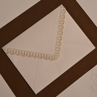 A close-up of cream-colored paper envelopes arranged in a geometric pattern with dark brown borders. One envelope is shown open, revealing a decorative cream lace corner trim and an embossed floral motif near the bottom.