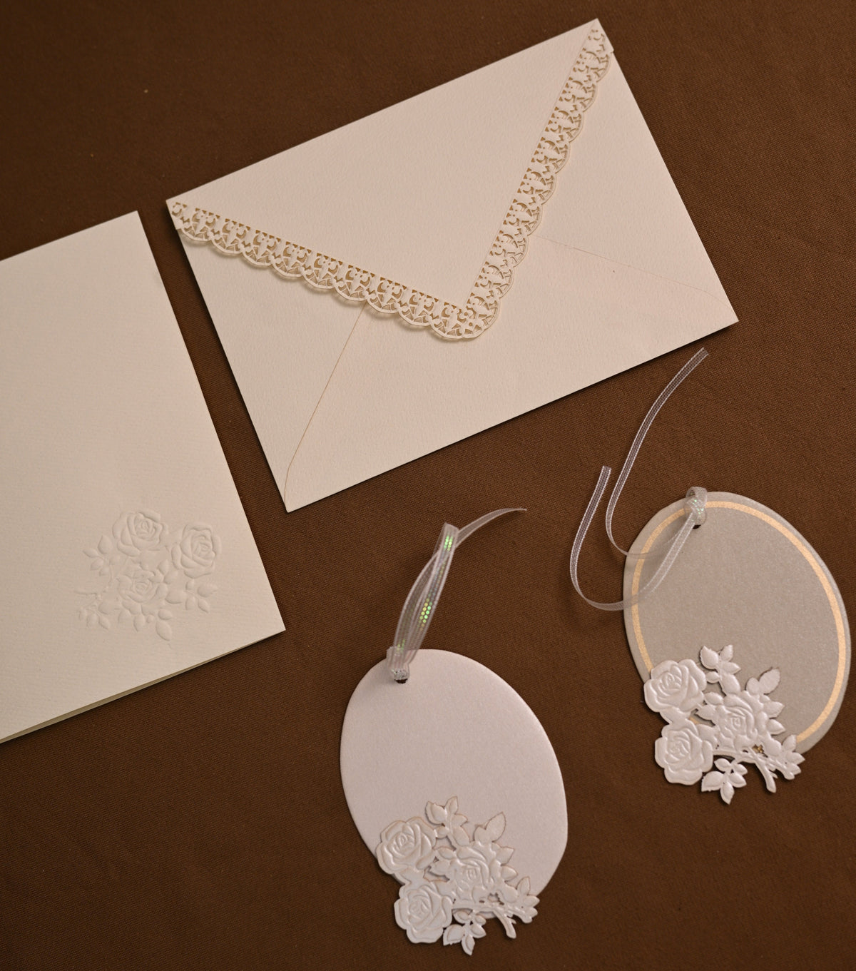 White wedding stationery set including an envelope with lace trim, a card with embossed floral detail, and two oval gift tags with lace embellishments, arranged on a dark brown background.
