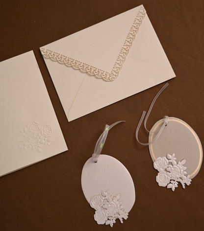 White wedding stationery set including an envelope with lace trim, a card with embossed floral detail, and two oval gift tags with lace embellishments, arranged on a dark brown background.