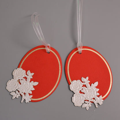 Two oval red gift tags with white rose cutout embellishments and clear hanging ribbons
