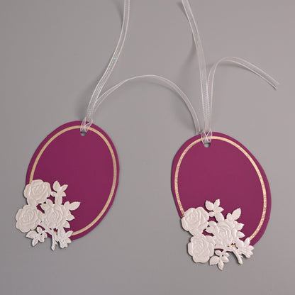 Two oval magenta gift tags with white floral cutouts and white ribbons, displayed on a neutral gray background.