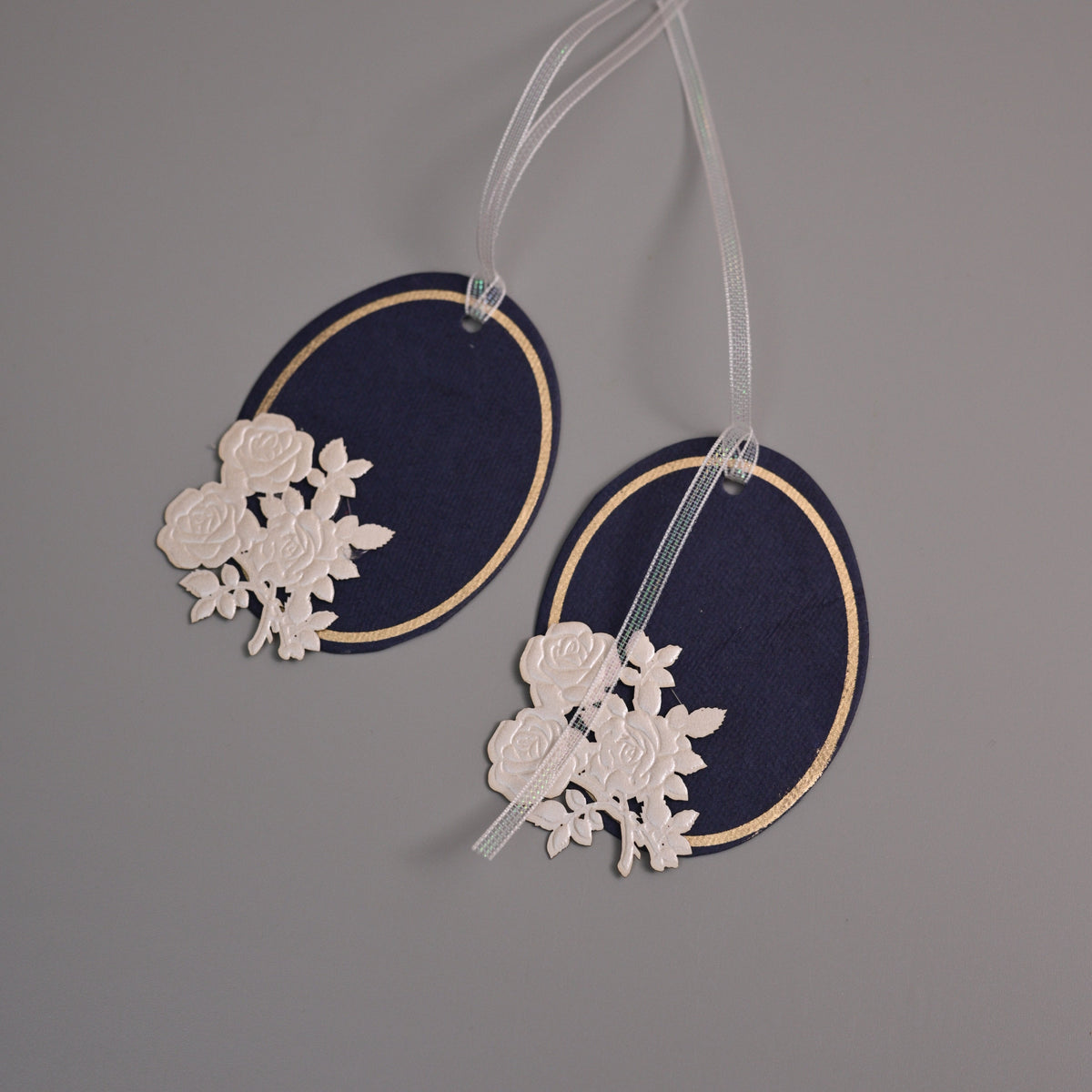 Two navy blue oval fabric tags with gold trim and white floral lace appliqué, hanging from clear ribbons against a plain gray background.
